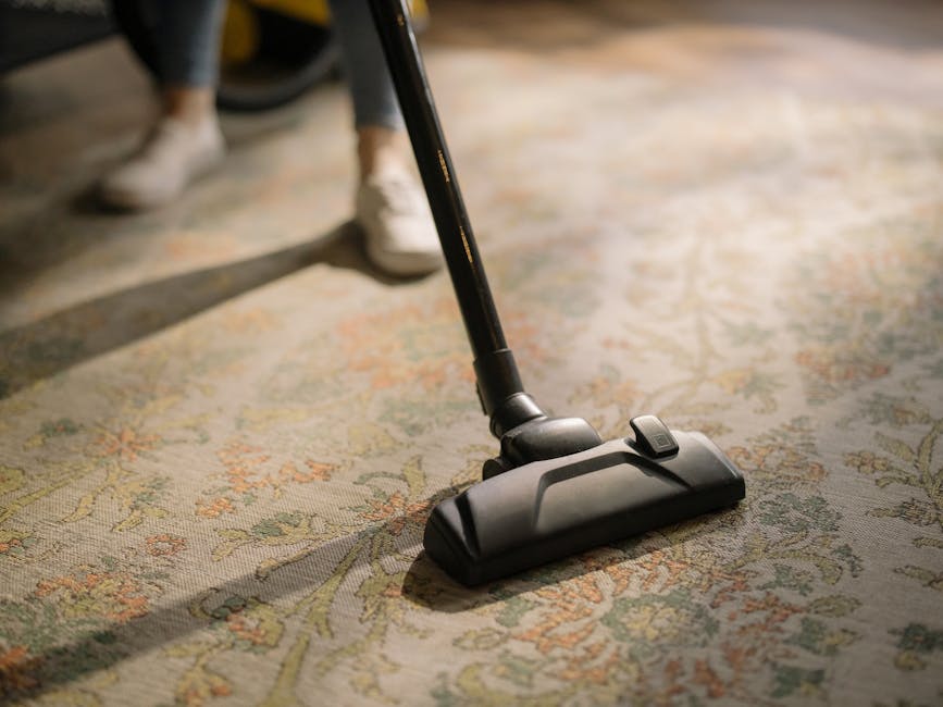 A person operating a vacuum cleaner on a patterned area rug with intricate floral designs in shades of beige, green, and orange, in a well-lit room. The vacuum cleaner's head is black, with a visible