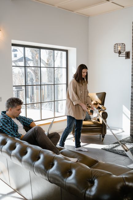 A woman is performing upholstery cleaning in a bright, modern living room with natural light streaming through large multi-pane windows. She is using a handheld vacuum cleaner or upholstery cleaner on