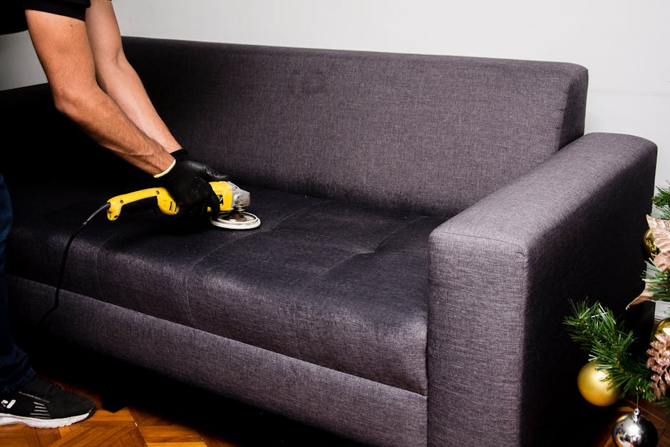 A woman is performing upholstery cleaning in a bright, modern living room with natural light streaming through large multi-pane windows. She is using a handheld vacuum cleaner or upholstery cleaner on